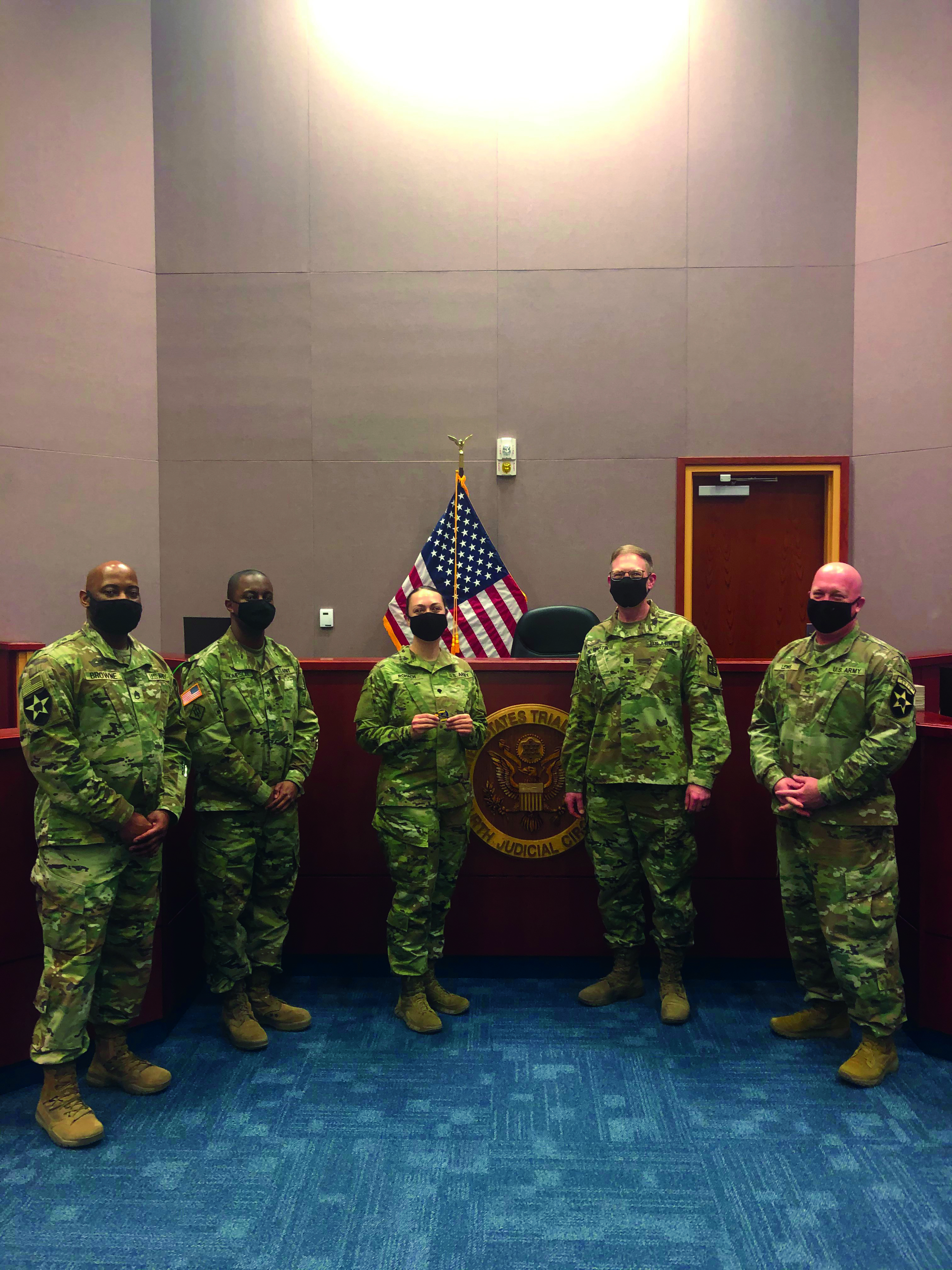 SPC Erica Rohach, 8th Army court reporter, proudly displays the U.S. Army Trial Judiciary coin she received for her recent success as the Distinguished Honor Graduate for the 64th Basic Court Reporter Course. The supporting team for the Korea/Japan Trial Judiciary, from L to R: SSG Browne, SSG Thompson, SPC Rohach, LTC Martin, and SFC Lewis. 